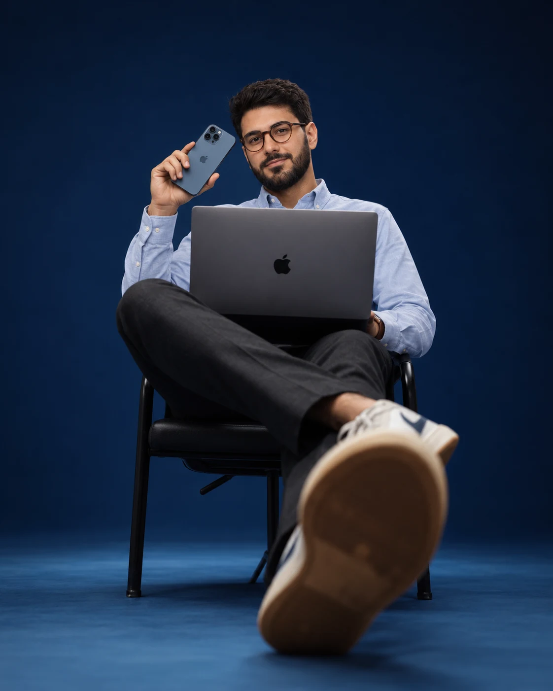 Gabriel Espinheira sitting on a chair in a navy photo studio, legs crossed toward the camera, a MacBook Pro on his lap and an iPhone raised beside his face.