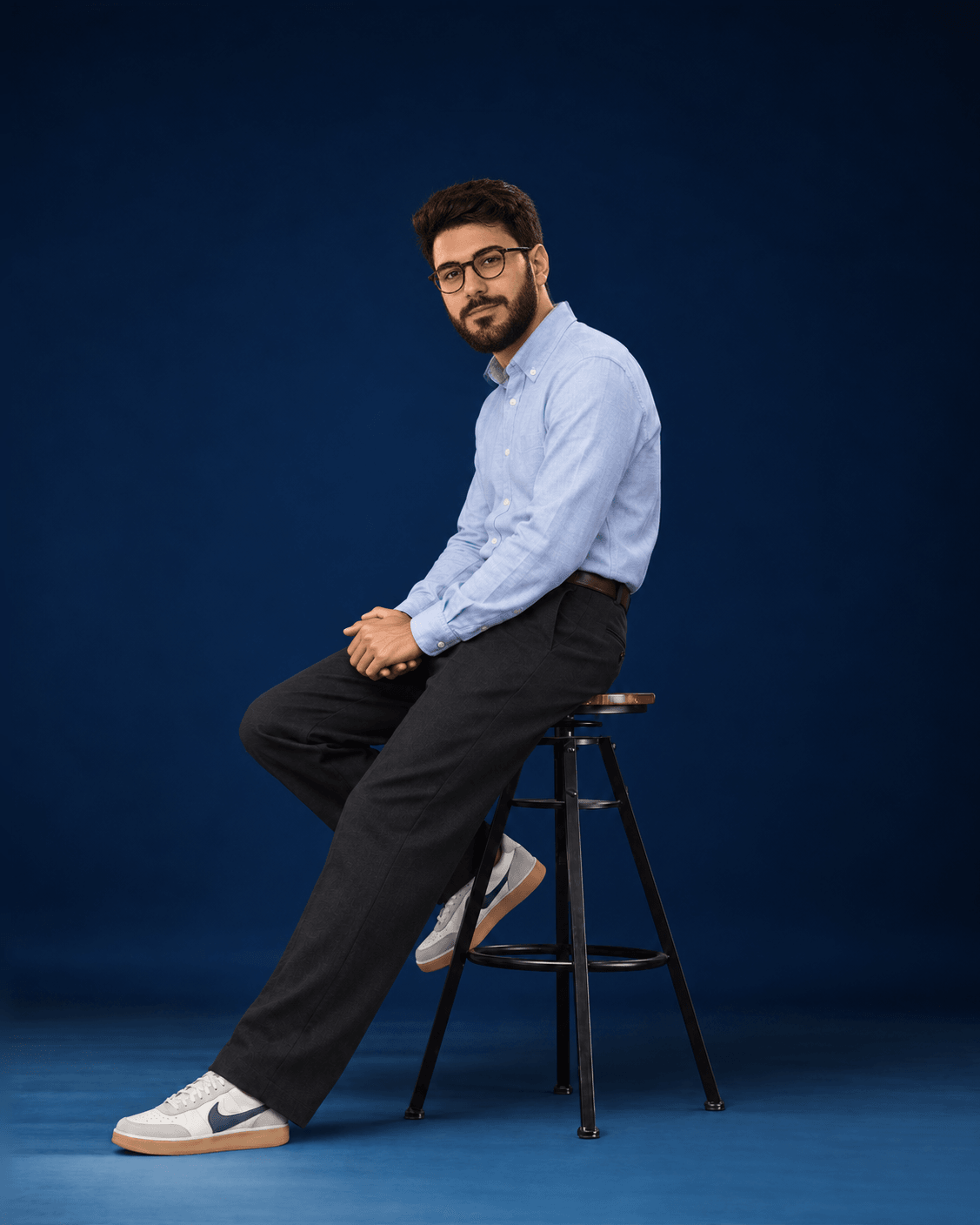 Gabriel Espinheira sitting on a stool in a navy photo studio, wearing a light blue shirt and dark trousers.